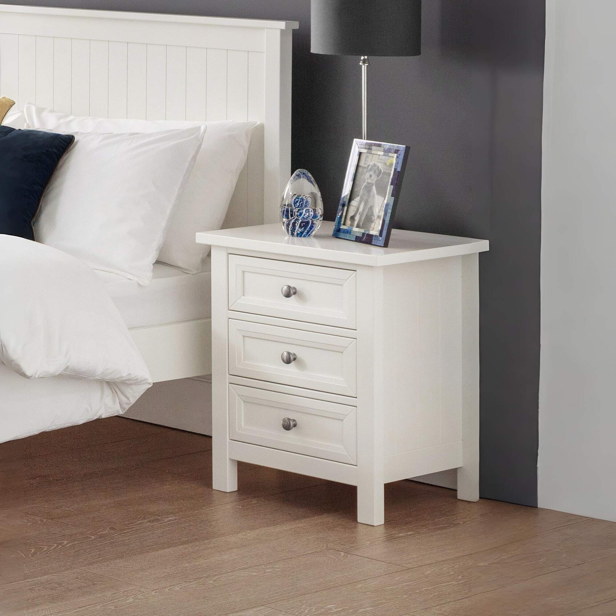 White nightstand with 3 drawers next to a bed with white bedding and a white headboard against a dark grey wall.